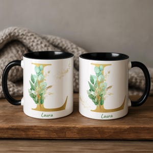 May include: Two white ceramic mugs with black handles and interiors. Each mug features a large gold letter L decorated with green eucalyptus leaves and gold accents. The name Laura is printed in green script below the letter on both mugs.