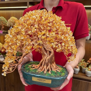 Pode incluir: Uma escultura decorativa de árvore bonsai com um tronco e ramos de fio de cobre grosso trançado, adornados com numerosas pequenas lascas de cristal citrino amarelo translúcido. A árvore está num vaso de cerâmica azul-petróleo escuro.