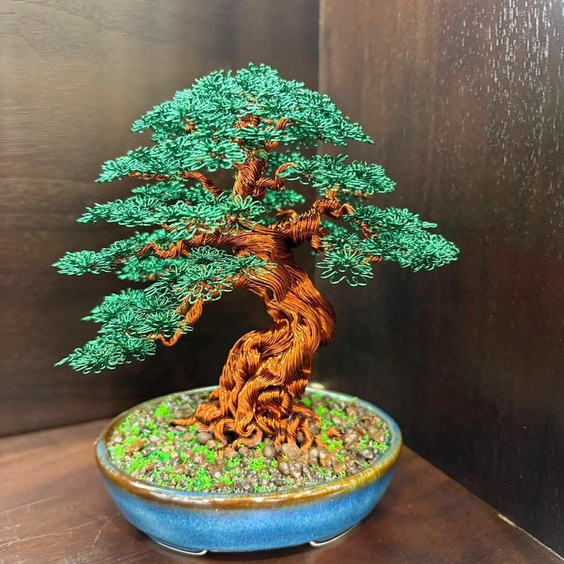 May include: A handcrafted wire bonsai tree sculpture. The tree features a twisted copper wire trunk and branches, with green wire foliage. It sits in a blue ceramic pot filled with small stones and moss. The bonsai is a decorative art piece.