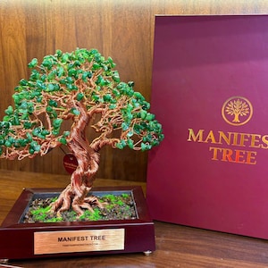 Puede incluir: Una escultura artesanal de árbol bonsái con un tronco y ramas de alambre de cobre, adornada con hojas de piedras preciosas verdes. El árbol está en una maceta de madera con musgo. Una caja burdeos con texto dorado "MANIFEST TREE" está en el fondo.