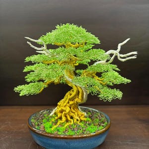 May include: A handcrafted bonsai tree sculpture. The tree features a twisted trunk and branches made of gold-colored wire, with bright green wire leaves. It sits in a blue ceramic pot filled with green moss and small rocks. The bonsai is a decorative item.