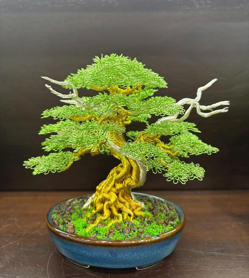 May include: A handcrafted bonsai tree sculpture. The tree features a twisted trunk and branches made of gold-colored wire, with bright green wire leaves. It sits in a blue ceramic pot filled with green moss and small rocks. The bonsai is a decorative item.