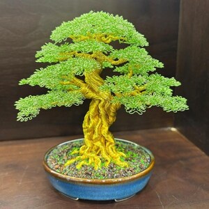 May include: A handcrafted bonsai tree sculpture. The tree features a twisted, golden-yellow trunk and branches, with vibrant green foliage. It is set in a blue ceramic pot filled with soil and small stones. The bonsai is a decorative piece.