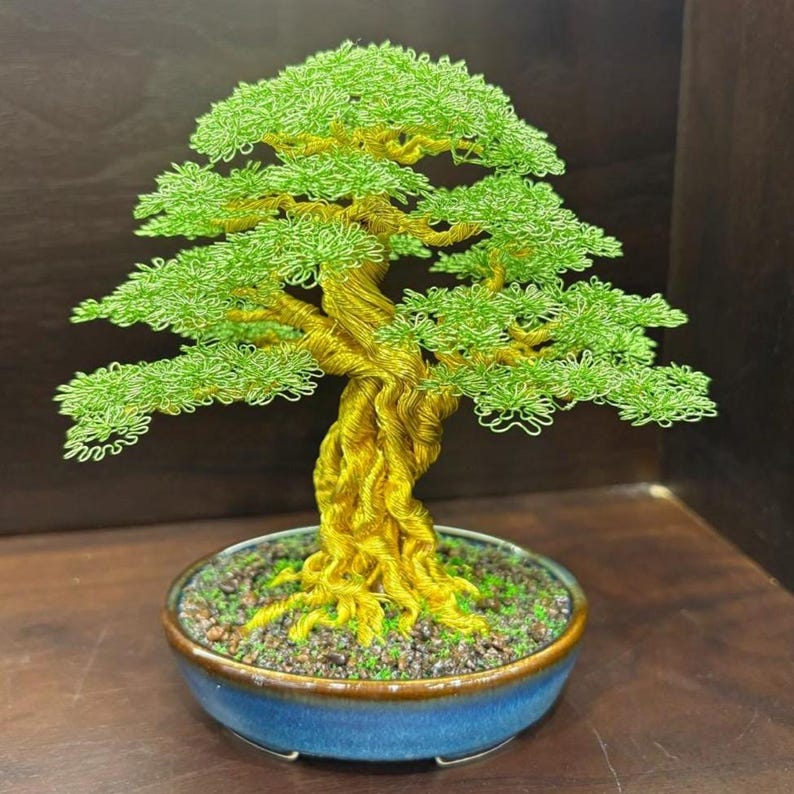 May include: A handcrafted bonsai tree sculpture. The tree features a twisted, golden-yellow trunk and branches, with vibrant green foliage. It is set in a blue ceramic pot filled with soil and small stones. The bonsai is a decorative piece.