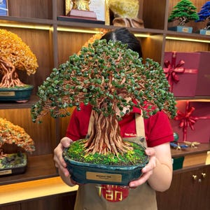 Pode incluir: Uma escultura decorativa de árvore bonsai com um tronco e ramos de cor cobre texturizados, adornados com numerosas pequenas folhas de pedra verde. A árvore está plantada num vaso de cerâmica oval verde-escuro com uma placa dourada com a inscrição MANIFEST TREE.