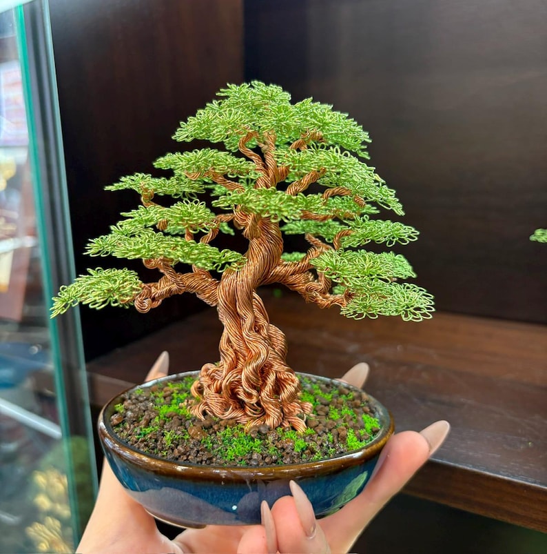 May include: A handcrafted bonsai tree sculpture made of copper wire, with green wire leaves. The tree is planted in a blue ceramic pot filled with soil and moss. The trunk and branches are intricately twisted, creating a detailed miniature tree.