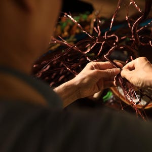 May include: Close-up of hands working on a copper wire sculpture. The sculpture has a tree-like design with many thin, branching wires. The hands are carefully manipulating the wires, likely during the assembly or shaping process. The overall color palette is warm, with copper and brown tones.