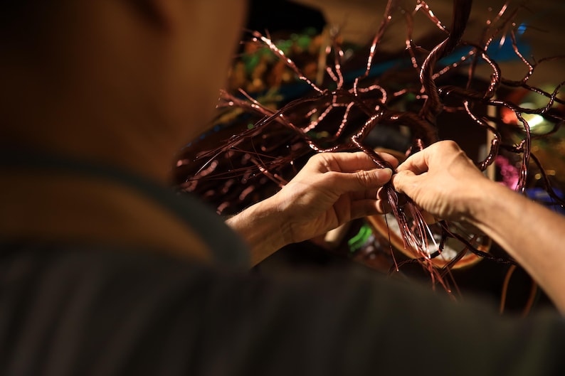 May include: Close-up of hands working on a copper wire sculpture. The sculpture has a tree-like design with many thin, branching wires. The hands are carefully manipulating the wires, likely during the assembly or shaping process. The overall color palette is warm, with copper and brown tones.