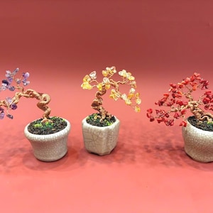 Op de afbeelding: Drie decoratieve bonsai-bomen met draad takken en gekleurde edelsteen bladeren. De bomen staan in witte keramische potten met een craquelé glazuur, tegen een rode achtergrond. De edelstenen zijn paars, geel en rood.