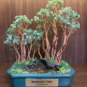 May include: A decorative sculpture featuring multiple copper wire trees with green stone leaves, set in a rectangular teal ceramic pot. The base includes a small pond with tiny fish figures and green moss. A metal plaque reads MANIFEST TREE TINHTANART.COM.