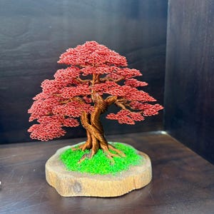 May include: A handcrafted wire bonsai tree sculpture with a red canopy and a twisted brown trunk. The tree sits on a wooden base with green moss. The sculpture is a decorative item.