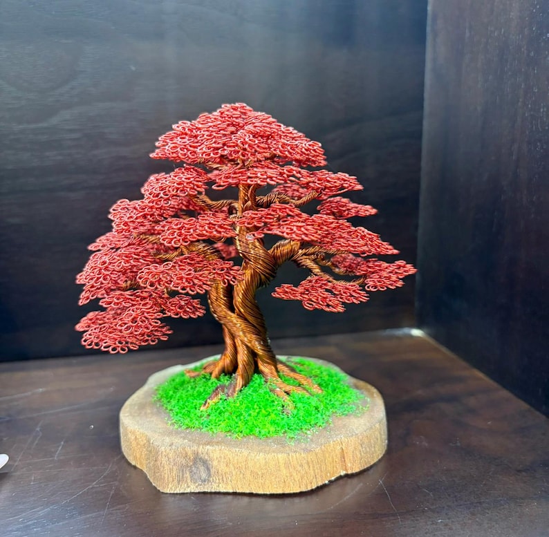 May include: A handcrafted wire bonsai tree sculpture with a red canopy and a twisted brown trunk. The tree sits on a wooden base with green moss. The sculpture is a decorative item.