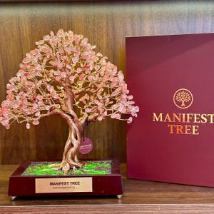 May include: A decorative Manifest Tree sculpture with a copper-colored trunk and branches, adorned with pink gemstone leaves. The tree sits in a dark wooden base with a green moss-like surface. A matching box with the words "MANIFEST TREE" is next to it.