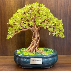 Könnte beinhalten: Ein dekorativer Bonsai-Baum mit einem gedrehten Kupferdrahtstamm und Ästen, verziert mit hellgrünen Kristallblättern. Der Baum steht in einem dunkelblauen Keramiktopf mit einer grünen, moosartigen Basis. Der Topf hat ein kleines rechteckiges Etikett mit der Aufschrift "MANIFEST TREE".
