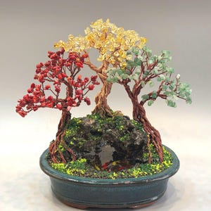 May include: A handcrafted bonsai tree sculpture with three trees, each with different colored gemstone leaves: red, yellow, and green. The trees are set in a dark green ceramic pot with a small pond and moss. The trunks are made of twisted copper wire.