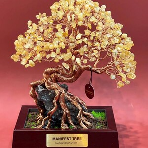 May include: A handcrafted copper wire tree sculpture with yellow gemstone leaves. The tree is mounted on a dark rock base within a wooden planter box. The sculpture has a tag that says "Metal Bonsai". The base has a gold label that says "MANIFEST TREE".