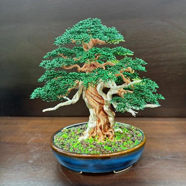 May include: A handcrafted bonsai tree sculpture. The tree features a twisted copper trunk, green wire foliage, and white branches. It is planted in a blue ceramic pot with a mossy base. The bonsai is a miniature representation of a mature tree.