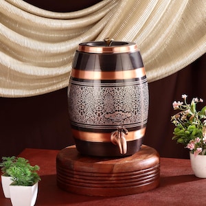 May include: A decorative, barrel-shaped beverage dispenser with a copper spigot. The dispenser features a black and copper finish with an ornate, etched design. It sits on a round, wooden base, and has a gold-colored handle on top.