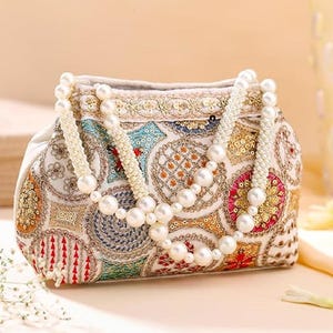 May include: An ornate, off-white clutch with intricate embroidery in various colors, including red, blue, and gold. A beaded pearl necklace drapes over the bag. The bag features a decorative pattern and a soft, neutral background.