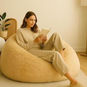 Könnte beinhalten: Eine Person sitzt auf einem großen, flauschigen, beigefarbenen Sitzsack und liest ein Buch. Sie trägt einen passenden beigefarbenen Pullover und eine Hose. Der Stuhl steht auf einem hellbeigen Teppich, mit einer Pflanze und einem Korbstuhl im Hintergrund.