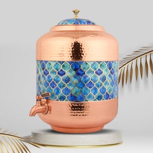 Puede incluir: Un dispensador de agua de cobre con acabado martillado. El dispensador presenta una banda de azulejos de mosaico azules y turquesas con un contorno dorado. Tiene un grifo de cobre y se asienta sobre una base de mármol blanco.
