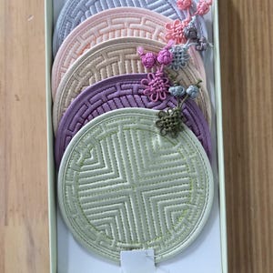 May include: A set of six round, embroidered coasters in a box. The coasters are in shades of gray, pink, beige, purple, and green, each with a geometric design. Decorative floral accents adorn the top of each coaster.