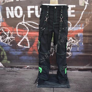 Op de afbeelding: Zwarte broek met groene accenten, bretels en metalen hardware. De broek heeft groene schedelontwerpen op de onderbenen en wordt gepresenteerd tegen een met graffiti bedekte muur. De broek heeft meerdere zakken en decoratieve stiksels.