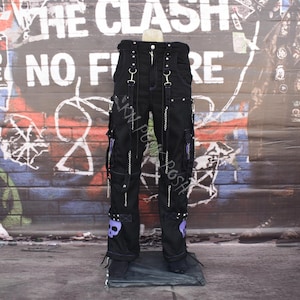 Op de afbeelding: Zwarte cargobroek met meerdere zakken, ritsen en zilverkleurige hardware. De broek heeft paarse schedelaccenten en riemen. De achtergrond bevat graffiti en posters, wat wijst op een punk- of alternatieve stijl.