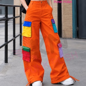 May include: Bright orange wide-leg cargo trousers featuring multiple utility pockets in contrasting colours including blue, yellow, teal, purple, green, and red. The trousers have a high waist, a button closure, and adjustable drawstring ties at the hem.