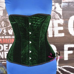May include: A dark green velvet corset with a structured silhouette. The corset features a series of silver-colored buttons down the front and a black trim along the edges. The garment is displayed against a backdrop of a blue top and a graffiti-style wall.