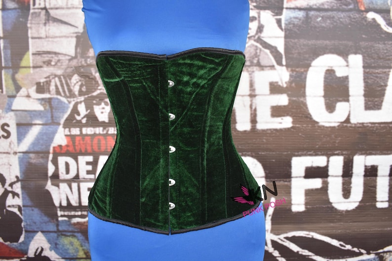 May include: A dark green velvet corset with a structured silhouette. The corset features a series of silver-colored buttons down the front and a black trim along the edges. The garment is displayed against a backdrop of a blue top and a graffiti-style wall.