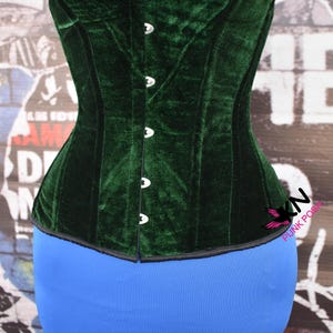 May include: A dark green velvet corset with a structured design. The corset has a row of silver-colored clasps down the front and a black trim along the edges. The garment is displayed on a blue form.