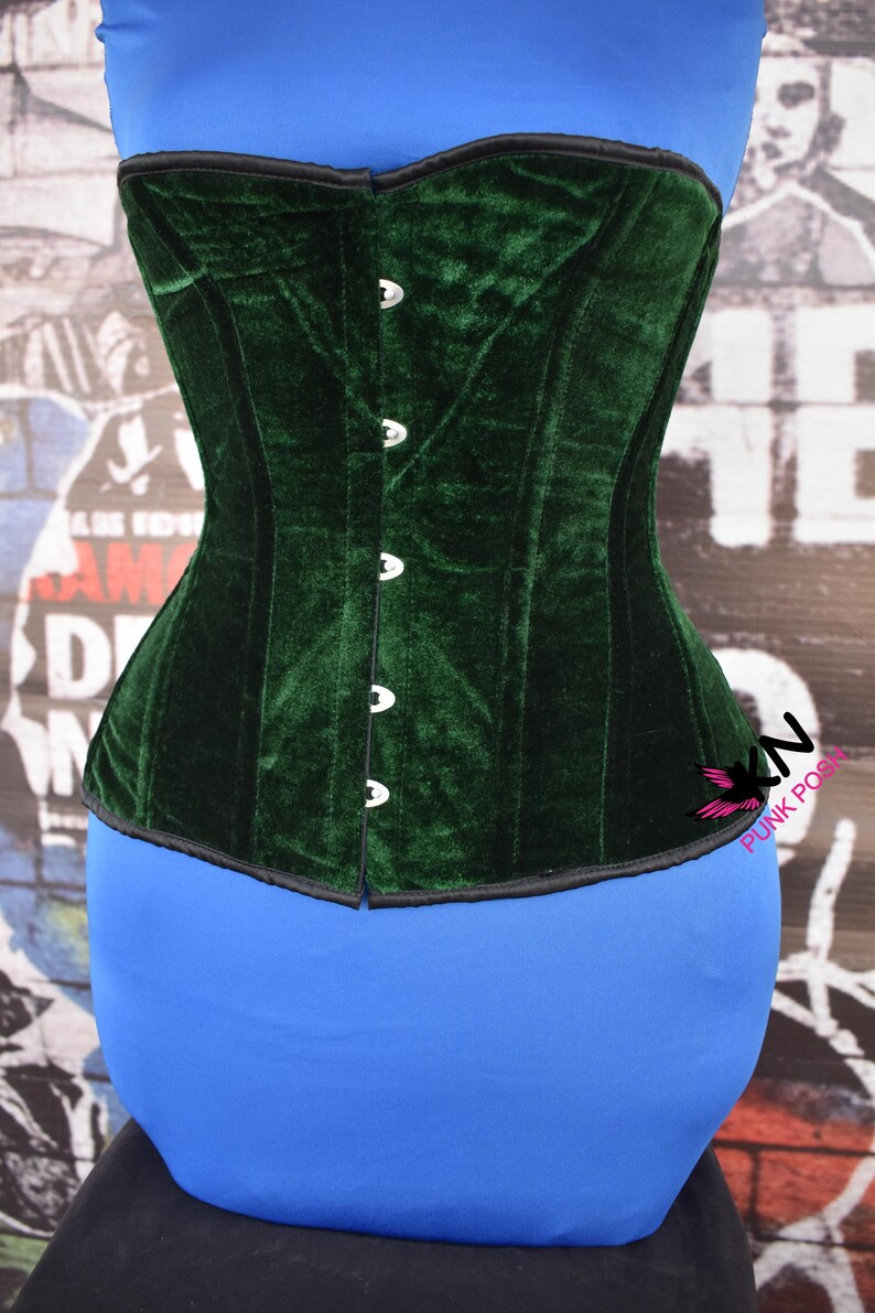 May include: A dark green velvet corset with a structured design. The corset has a row of silver-colored clasps down the front and a black trim along the edges. The garment is displayed on a blue form.