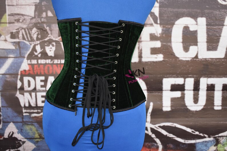May include: A dark green velvet corset with black lacing. The corset is laced up the back and has a structured design. The background is a graffiti-style wall with faded text.