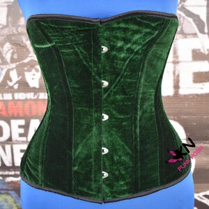 May include: A dark green velvet corset with a black trim. The corset has a series of silver-colored clasps down the front and is displayed on a blue form. The corset is a piece of clothing.