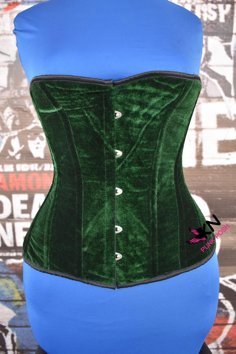 May include: A dark green velvet corset with a black trim. The corset has a series of silver-colored clasps down the front and is displayed on a blue form. The corset is a piece of clothing.