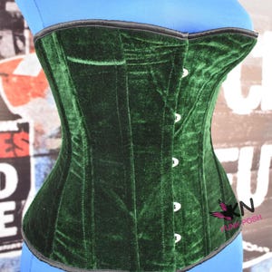 May include: A dark green velvet corset with a black trim and white buttons. The corset is a structured garment with vertical seams and a curved shape. The corset is displayed on a blue form.