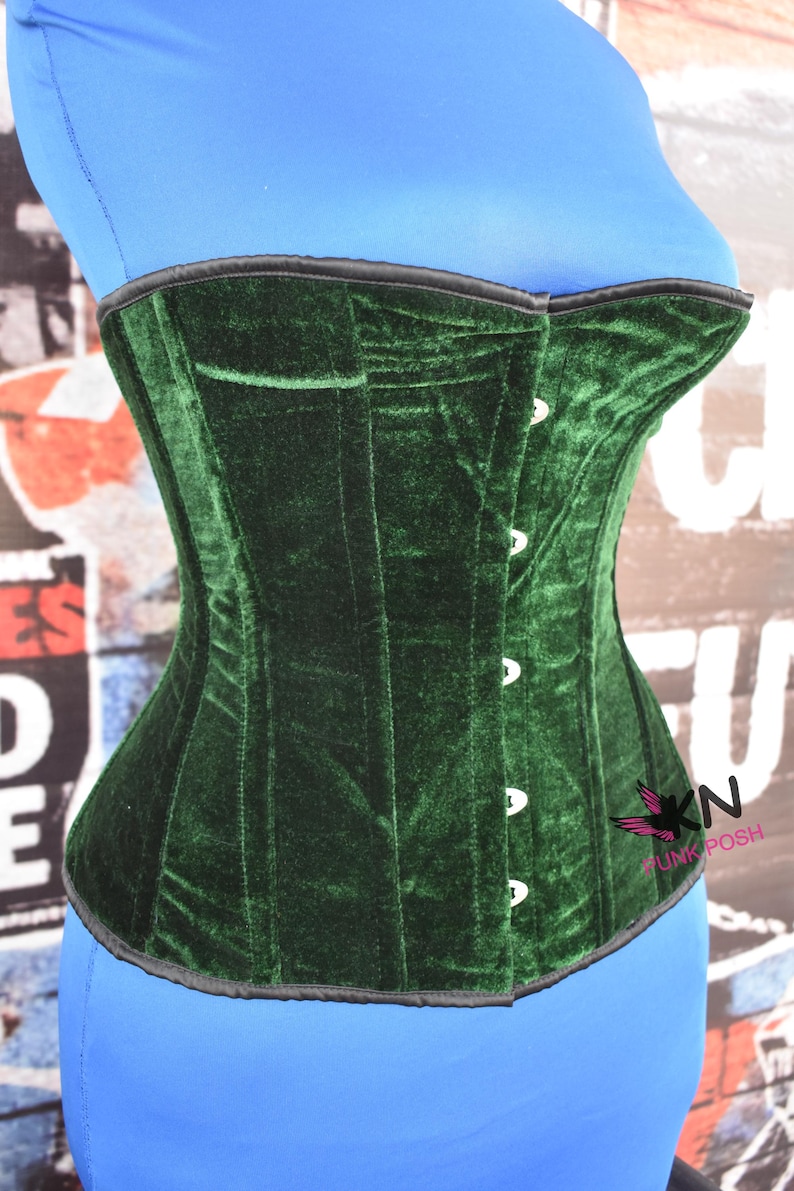 May include: A dark green velvet corset with a black trim and white buttons. The corset is a structured garment with vertical seams and a curved shape. The corset is displayed on a blue form.