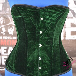 May include: A dark green velvet corset with a structured design. The corset features a row of silver-colored clasps down the front and a black trim along the top and bottom edges. The corset is displayed on a blue form.
