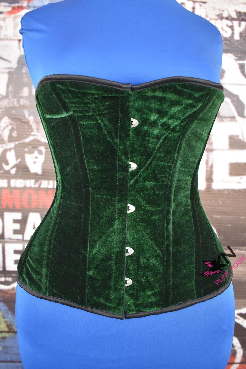 May include: A dark green velvet corset with a structured design. The corset features a row of silver-colored clasps down the front and a black trim along the top and bottom edges. The corset is displayed on a blue form.
