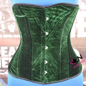 May include: A dark green velvet corset with a black trim. The corset has a structured design with vertical panels and is fastened with silver-colored clasps. The corset is displayed on a blue form, with the text "KN PUNK POSH" visible.