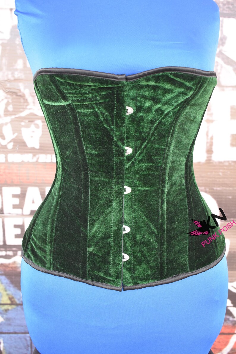 May include: A dark green velvet corset with a black trim. The corset has a structured design with vertical panels and is fastened with silver-colored clasps. The corset is displayed on a blue form, with the text "KN PUNK POSH" visible.