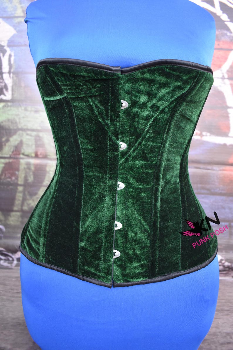 May include: A dark green velvet corset with a black trim and silver-colored clasps. The corset is displayed on a blue form. The brand logo "KN Punk Posh" is visible on the right side.