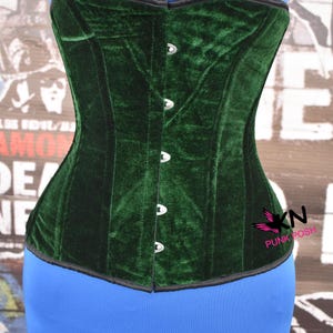 May include: A dark green velvet corset with a structured design. The corset features a black trim and silver-colored clasps down the front. The corset is displayed on a blue form. The logo "KN PUNK POSH" is visible on the right side.