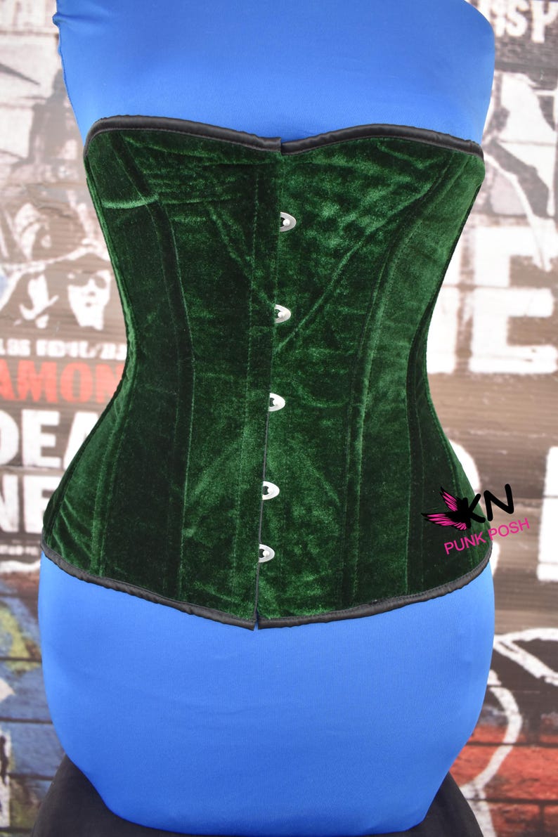 May include: A dark green velvet corset with a structured design. The corset features a black trim and silver-colored clasps down the front. The corset is displayed on a blue form. The logo "KN PUNK POSH" is visible on the right side.