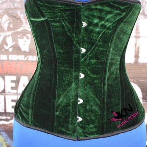 May include: A dark green velvet corset with a black trim and silver-colored clasps. The corset has a structured design and a slight curve at the waist. The logo "KN Punk Posh" is visible on the lower right side.