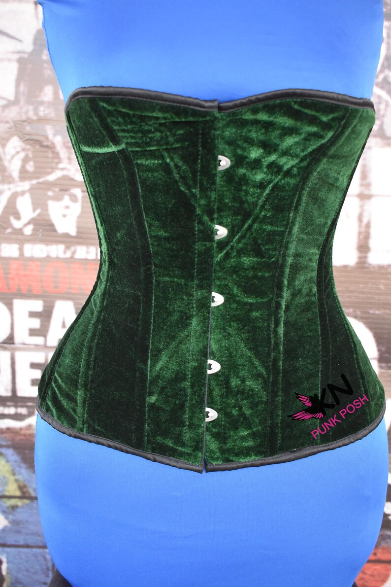 May include: A dark green velvet corset with a black trim and silver-colored clasps. The corset has a structured design and a slight curve at the waist. The logo "KN Punk Posh" is visible on the lower right side.
