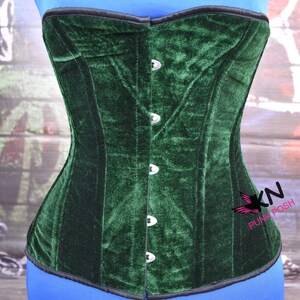 May include: A dark green velvet corset with a black trim and silver-colored clasps. The corset has a structured design and is displayed on a blue form. The corset is from the brand "KN Punk Posh".