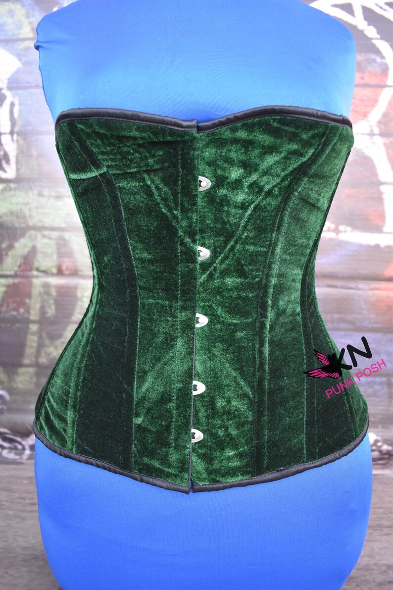 May include: A dark green velvet corset with a black trim and silver-colored clasps. The corset has a structured design and is displayed on a blue form. The corset is from the brand "KN Punk Posh".
