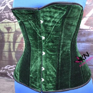 May include: A dark green velvet corset with a structured design. The corset features a row of silver-colored buttons down the front and black trim along the edges. The corset is displayed on a blue form.